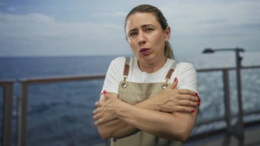 Tekne güvertesinde kollarını saran bir kadın bulutlu gökyüzünün altında at kuyruklu bir örtü içinde rahatsız;.