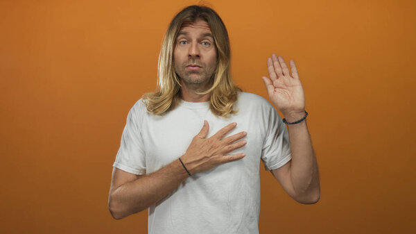 Man with long blond hair in white shirt raises right hand and places palm to chest in orange studio setting; sincerity.