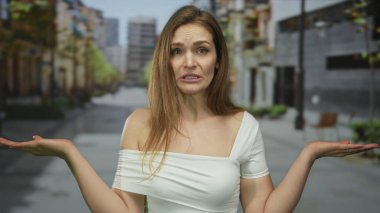 Beyaz omuzlu genç kadın, yüksek binaların arasında boş şehir caddesinde avuçlarını silkeliyor; belirsizlik.
