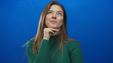 Eli çenesinde olan bir kadın mavi stüdyoda dik dik bakıyor yeşil balıkçı yaka kazak giyiyor..