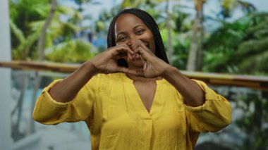 Sarı tişörtlü kıvırcık saçlı bir kadın havuz başında gülümserken elleriyle kalp şeklinde bir kalp yapıyor, açık havada neşeli bir tropikal titreşim yakalıyor..