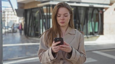 Güneşli şehir caddesinde elinde akıllı telefon tutan genç bir kadın bej renkli trençkotla yukarı bakıyor..