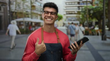 Önlüklü gülümseyen adam elinde tabletle şehir terası kafesinde arka planda insanlar ve binalar var.