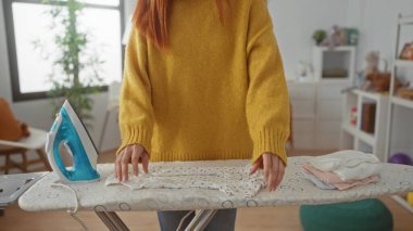 Konforlu ev çamaşırhanesinde kıyafetlerini ütüleyen, sarı kazak giyen, camdan içeri güneş ışığı süzülen ve temiz bir yaşam tarzını ön plana çıkaran bir kadın.