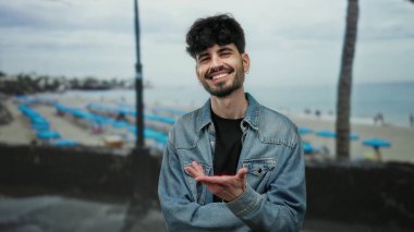 Kumsalda gülümseyen genç bir adam bulutlu bir gökyüzünün altında kot ceket giyiyor, deniz kenarında bir atmosferde boş zamanlarını ve mutluluğu yakalıyor..