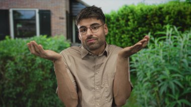 Bej gömlekli adam, yeşil çitin çerçevelediği girişte omuz silkiyor; iç belirsizlik.