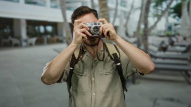 Elinde kamera olan beyaz sakallı genç bir adam şehir sokağında fotoğraf çekimi için bekliyor. Sırt çantası takıyor ve turist gibi görünüyor..