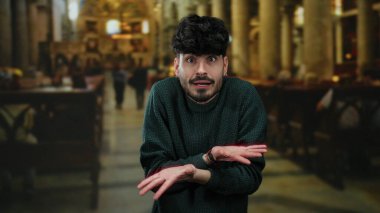 Tarihi kilise ortamında şaşkın bakışları olan genç bir adam, süveter giyiyor, kapalı bir arkaplanda duygularını sergiliyor..