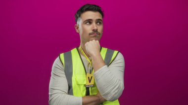 Yansıtıcı yelek giyen genç İspanyol adam, gönüllü rozeti ile izole edilmiş, pembe bir arka plana karşı düşünceli bir şekilde duruyor..