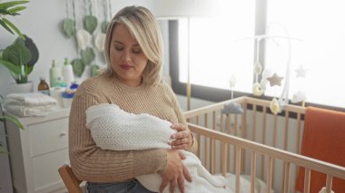 Genç sarışın kadın kundaklanmış bebeği binada tahta beşiğin yanında tutarken esniyor; yorgunluk tükenmişlik.
