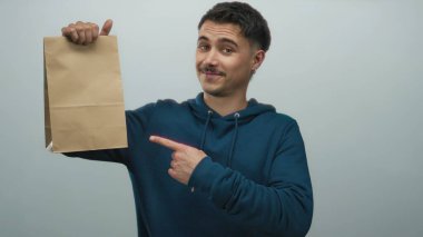 Mavi kapüşonlu ve gülümserken olumlu jestler yapan, izole edilmiş beyaz arka planda kağıt çanta tutan genç İspanyol adam..