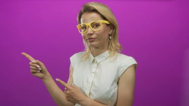 Sarı gözlüklü ve beyaz kolsuz bluzlu bir kadın mor bir stüdyoyu işaret ediyor..