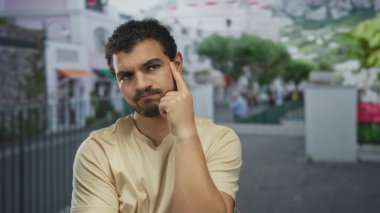 Çapraz kolları ve büzüşmüş dudakları olan genç adam gün ışığında beyaz bina cephesinin yanında duruyor. İnkar..