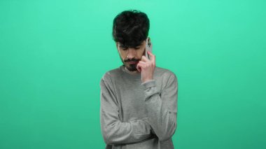 Gri kazaklı genç adam, kapalı gözlü, düşünceli bir yüz ifadesi gösteren izole edilmiş yeşil bir arka plana karşı akıllı telefondan konuşuyor..
