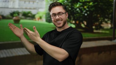 Parkta sakallı ve gözlüklü genç adam, güneşli bir günde yeşil arka planda bir şey sunan, neşeli bir yüz ifadesi gösteren elini uzatıyor..