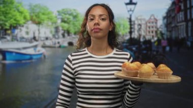 Woman holding a tray of muffins with fist raised and hand visible on a street in amsterdam; determination sharing.