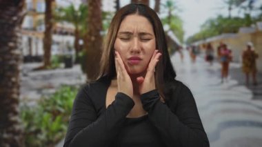 Genç İspanyol kadın gözleri kapalı ve elleri yanaklarında diş ağrısı için yeşil güneş ışığı sokağında; rahatlama.