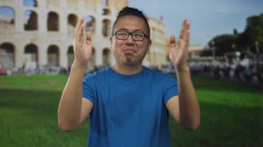 Elleri havada, Roma kolezyum binasında el kol hareketleri yapan, gözlüklü ve mavi tişörtlü, elleri antik kemerleri çerçeveleyen bir adam..