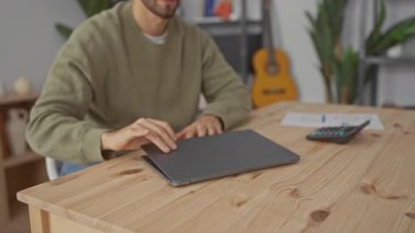 Genç adam rahat bir oturma odasında dizüstü bilgisayarla çalışıyor. Bitkiler ve gitarın bulunduğu bir odada. Yakınındaki ahşap masada hesap makinesi olan bir hesap tablosuna odaklanıyor..