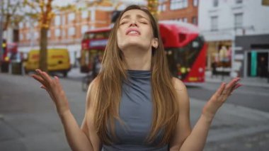 Kafkasyalı genç kadın ellerini kaldırıyor ve kırmızı otobüs ve tuğla binayla caddelerde gözlerini kapatıyor; hayal kırıklığı.