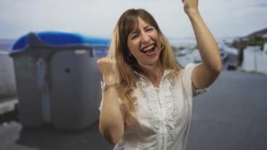 Bir şehir caddesinde geri dönüşüm konteynırının yanında yumruklarını sıkmış bir kadın, kaldırılmış kollarla gülüyor ve tezahürat yapıyor; sevinç kutlaması.
