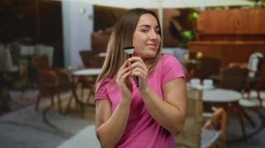 Elinde makyaj fırçasıyla gülümseyen, pembe gömlek giyen restoran terasında oturan şehir sokaklarında güzellik ve rahatlık sergileyen bir kadın..