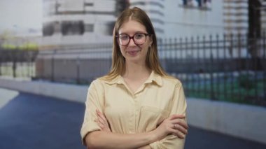 Şehir caddesinde tarihi binanın yanında kolları çaprazlanmış gülümseyen bej düğmeli bir kadın..