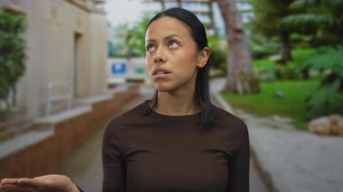 Siyah saçlı genç bir kadın yeşil bir park ortamında yukarı bakıyor, düşünceli ve sakin görünüyor, açık havada kahverengi bir bluz giyiyor yemyeşil ve doğa ile çevrili..