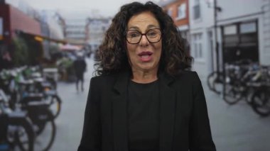 Kıvırcık saçlı ve gözlüklü İspanyol kadın arka planda bulanık bisiklet ve binalarla çevrili kalabalık bir şehir sokağında şaşkınlığını dile getiriyor..