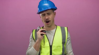 Yansıtıcı yelek giymiş genç İspanyol bir adam elinde bir matkap tutuyor ve izole edilmiş pembe bir arka plana yaslanıyor. İnşaat sırasında yorgunluk hissi veriyor..