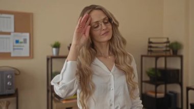 Ofis ortamında gözlüklü genç sarışın kadın saçlarını düzeltirken saksıların önünde çiçek ve ofis belgeleriyle duruyor..