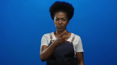 Önlük takan genç bir kadın, izole edilmiş mavi arkaplan karşısında şakacı hareketler sergiliyor.