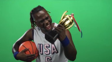 Elinde altın bir kupa ve basketbol topu tutan, ABD forması ve bileklikleriyle stüdyoda zafer kutlamaları yaparken ağzı açık bağıran bir adam..