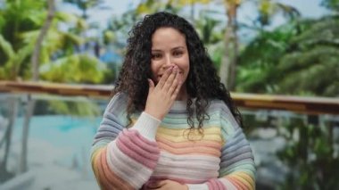 Güneşli bir günde, yemyeşil ve canlı tropikal bitkilerle çevrili bir otelde havuz kenarında renkli bir süveter içinde kıvırcık saçlarıyla gülümseyen genç İspanyol kadın..