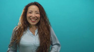 Dişleriyle gülümseyen, uzun kıvırcık saçlı, açık mavi tişörtlü, deniz mavisi stüdyosunda kameraya bakan bir kadın..