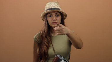 Hasır şapkalı bir kadın kahverengi bir arka planın önünde özgüvenli bir turist havası yaratıyor ve boynunda klasik bir kamera asılı duruyor..