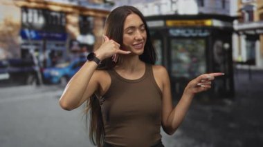 Genç İspanyol kadın gülümsüyor, işaret işareti yapıyor ve caddede kahverengi bir tişört giyerken işaret ediyor; dostça bir davet.