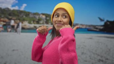 Parlak pembe kazaklı genç İspanyol kadın deniz kenarında neşeyle gülümseyen sarı bir bere takan parlak mavi gökyüzü ile kumsalda geriye doğru hareket ediyor.