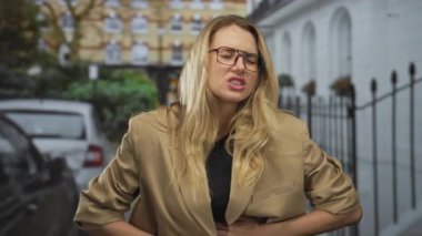 Sarışın bir kadın, şehir sokaklarında karın ağrısı çekiyor. Gözlük ve bej ceket takıyor. Arabalara ve binalara yaslanırken rahatsız olduğunu söylüyor..