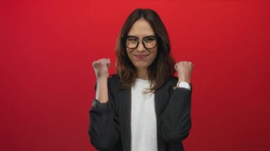 Kadın zaferi canlı kırmızı arka plana karşı yumruklarını kaldırarak kutluyor. Gözlüklü ve çizgili ceketli. Başarıyı ve mutluluğu simgeliyor..