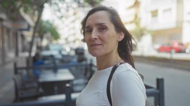 Woman smiling outdoors on a sunny street wearing white shirt with parked cars and buildings in the background.
