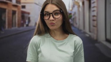 Gözlüklü kız gülümsüyor ve taş binalarla dar bir şehir sokağında çerçeveyi düzeltirken dilini dışarı çıkarıyor; oyuncu tavır..