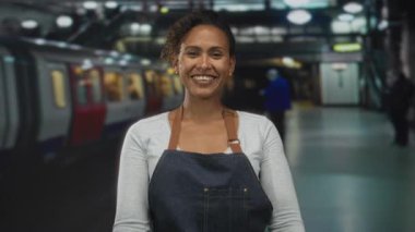 Kot önlüklü kadın gülümsüyor ve elleri açık bir tren istasyonunda, demiryolu koridorundaki kameraya bakıyor; hoş geldiniz..