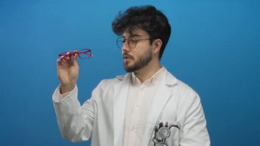 Sakallı, mavi arka planda kırmızı gözlük takan, steteskoplu beyaz laboratuvar önlüğü giyen, düşünceli bir yüz ifadesi sergileyen İspanyol genç adam..