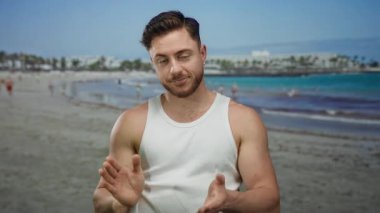 Sahilde sakallı genç bir adam arka planda insanlarla el çırpıyor açık gökyüzünün altında kumsalın tadını çıkarıyor.