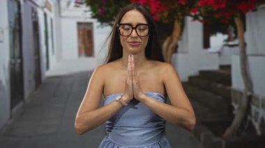 Elleri birbirine bastırılmış genç İspanyol kadın güneş ışığı altındaki dar bir sokakta gözlük ve askısız elbise giyerek dua ediyor. Huzur meditasyon farkındalığı..