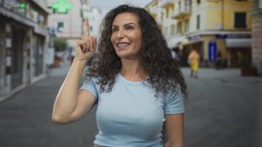 Açık mavi tişörtlü gülümseyen genç İspanyol kadın kalabalık şehir caddesinde parmağını yukarı kaldırıyor. İyimserlik.