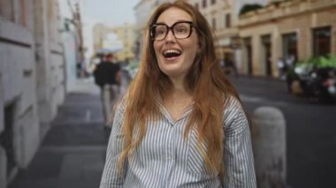 Genç beyaz kadın gözlük takıyor ve kalabalık bir şehir caddesinde dururken geniş bir gülümseme sergiliyor; mutluluk iyimserliği.