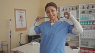Mavi önlüklü genç kadın bir klinik odasında steteskop tutuyor. Arka planda tıbbi posterler ve malzemelerle güvenle gülümsüyor..