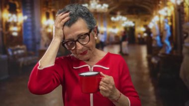 Kırmızılı yaşlı kadın elinde parlak bir kilisenin içinde derin derin düşünen bir kahve fincanı tutuyor, avizeler ve heykeller sıcak bir ortam oluşturuyor..